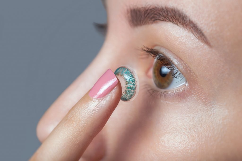 woman holds a blue colored contact lens on her finger. 
