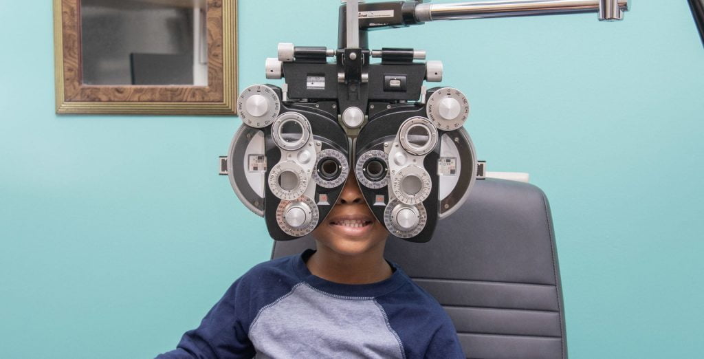 boy sits in eye exam room with phoropter over his eyes