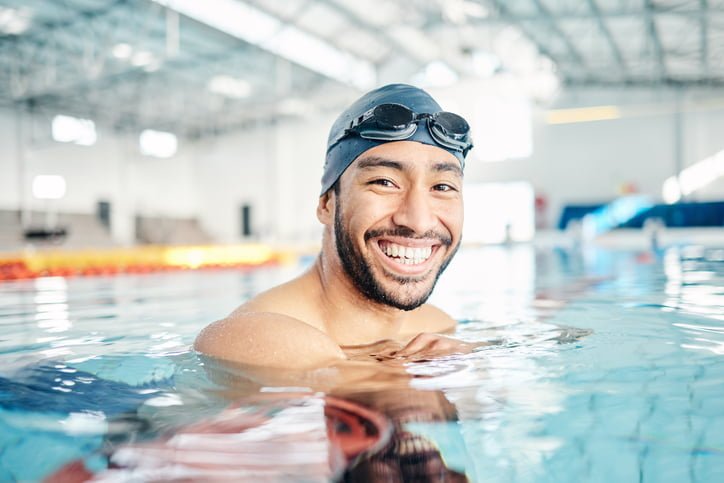 Can you wear contacts in the pool? Studies show it's not safe to swim with contact lenses. Image shows man in pool with goggles on his head, smiling. 